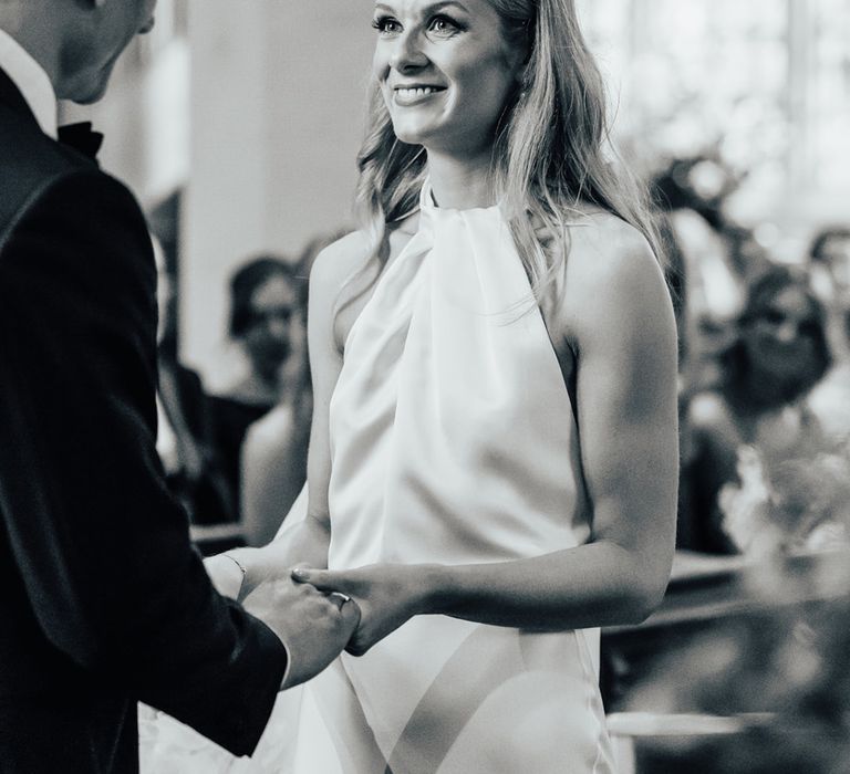 Bride in halterneck Halfpenny London wedding dress with floral headpiece holding hands with groom at the altar during church wedding ceremony