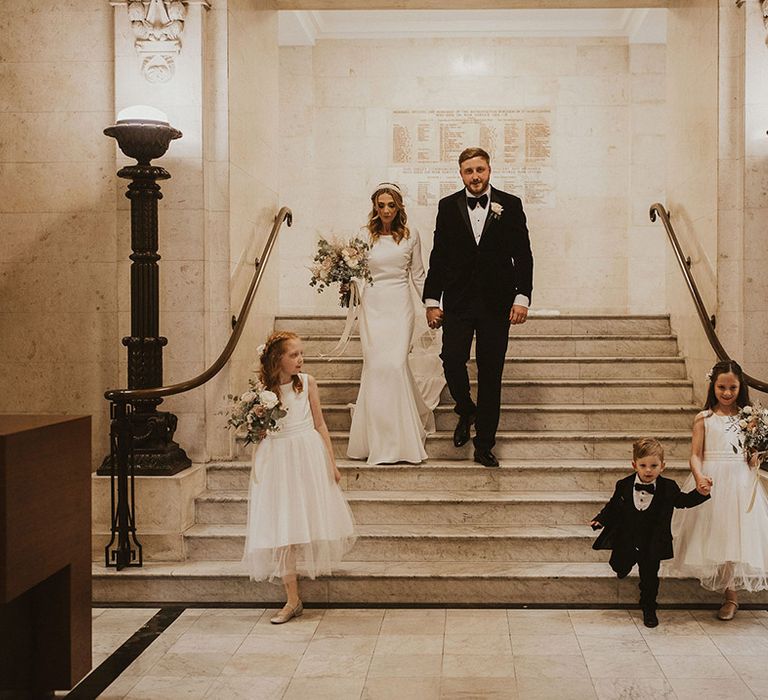 Bride in a Pronovias wedding dress with long sleeves and groom in a tuxedo walking down the steps at Old Marylebone Town Hall with their children 