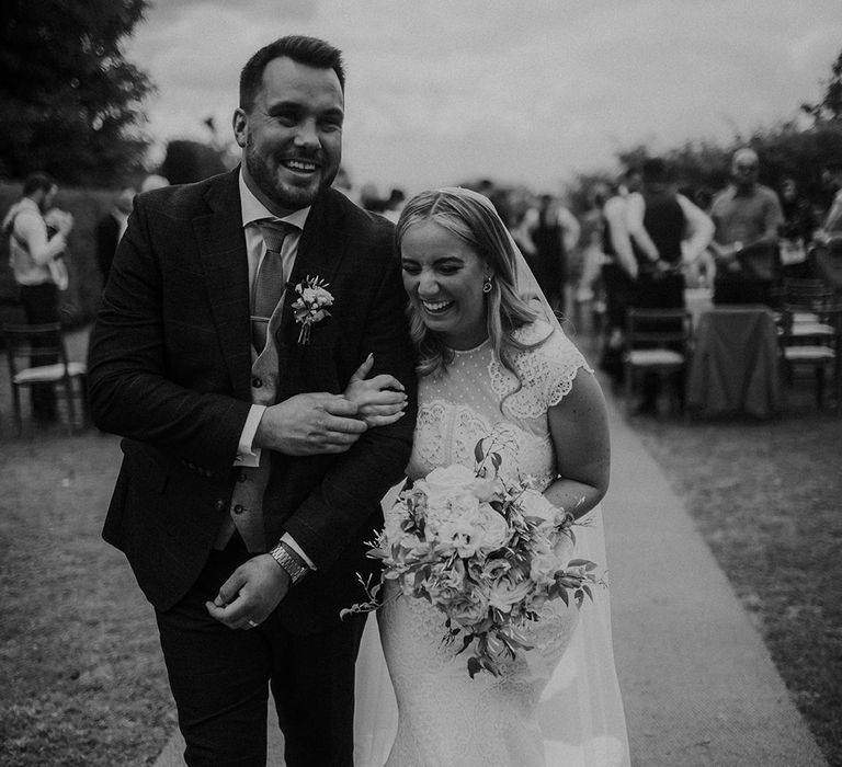 Black and white portrait of the bride in a lace wedding dress arm in arm with her groom walking up the aisle 