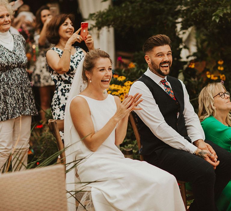 Bride in a slip wedding dress and pinned up hair and groom in a navy waistcoat and floral tie sitting on wooden chairs laughing during the speeches 