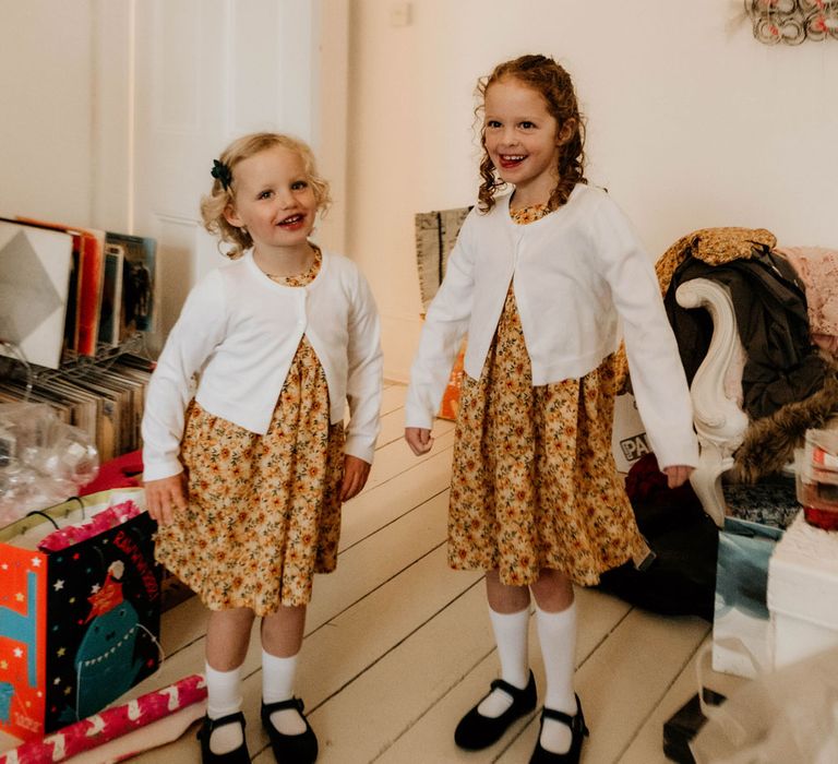 Two flower girls in sunflower dresses, white cardigans and long white socks stands together in lounge before wedding