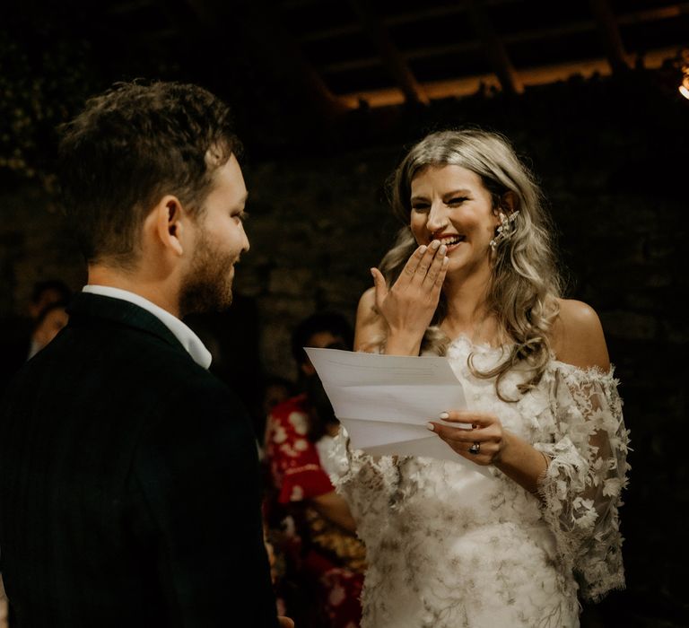 Bride with curled hair and wedding dress with applique flowers laughs whilst reading her vows during bespoke ceremony