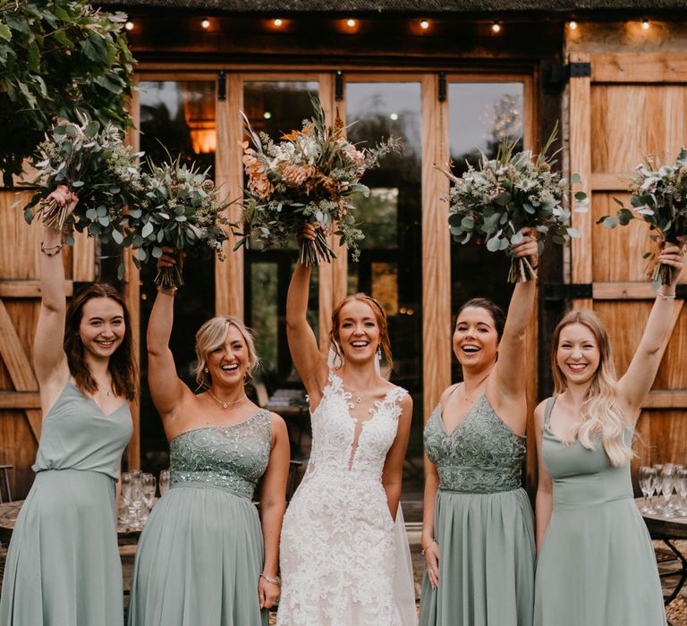 Bride in deep v lace wedding dress with tulle skirt and bridesmaids in sage green bridesmaid dresses hold up mixed wedding bouquets outside barn after wedding ceremony