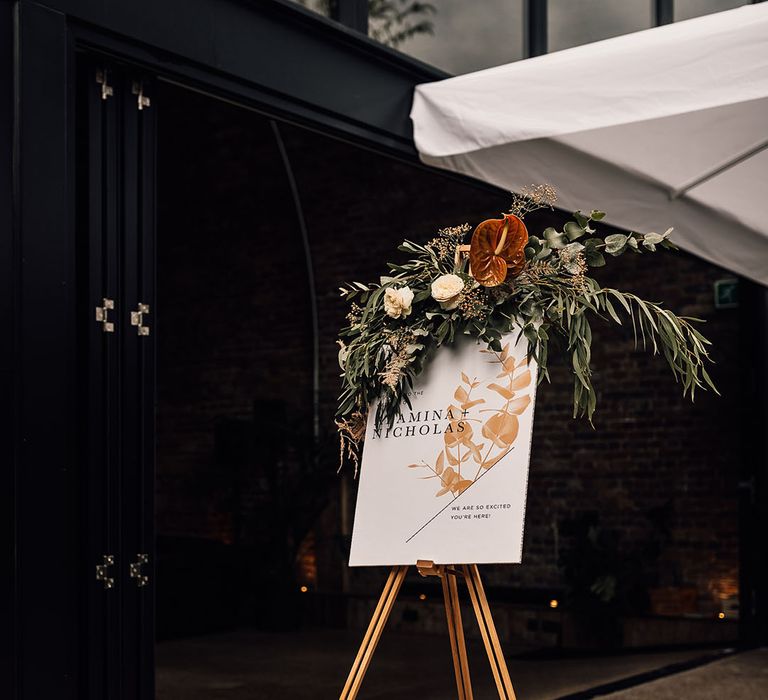 Wedding welcome sign with floral design and orange anthurium and green foliage decor on the wooden easel 