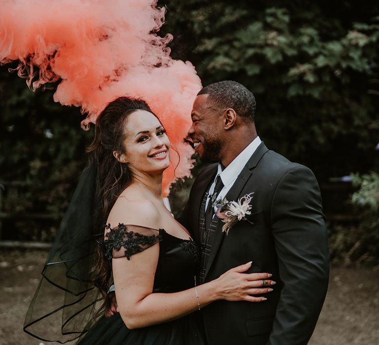 Bride & groom hold pink smoke bomb as they embrace after wedding ceremony