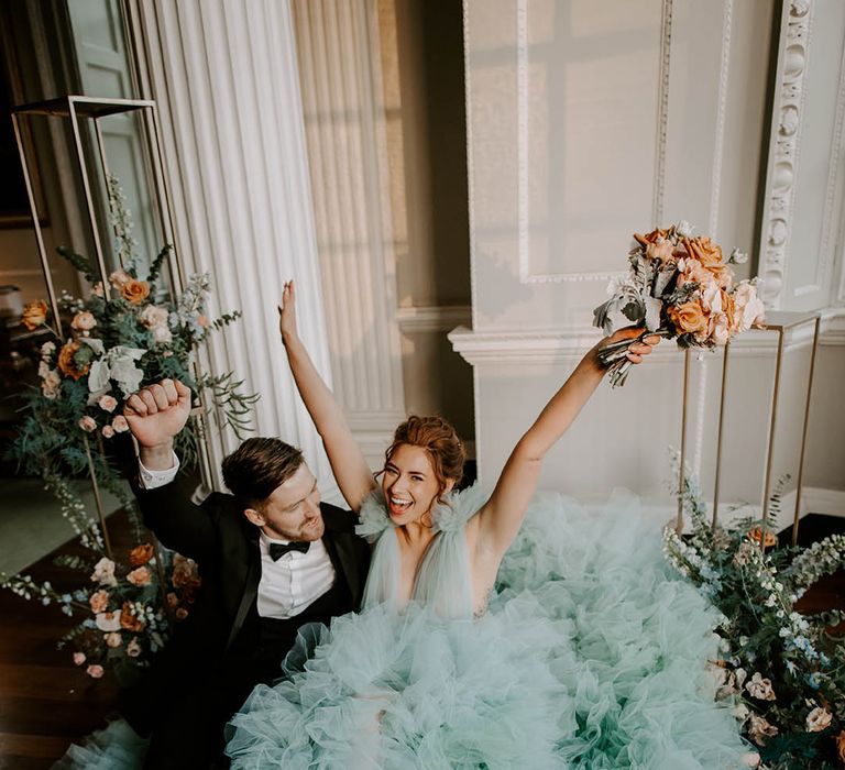 Bride in a light blue wedding dress and bow front shoes holding her cafe au late wedding bouquet in the air as she sits with her groom in a tuxedo 