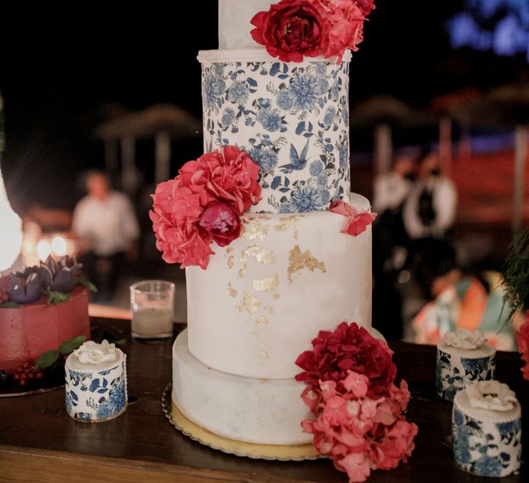 Four tier wedding cake complete with white frosting, blue floral design, gold leaf and bright red floral decor 