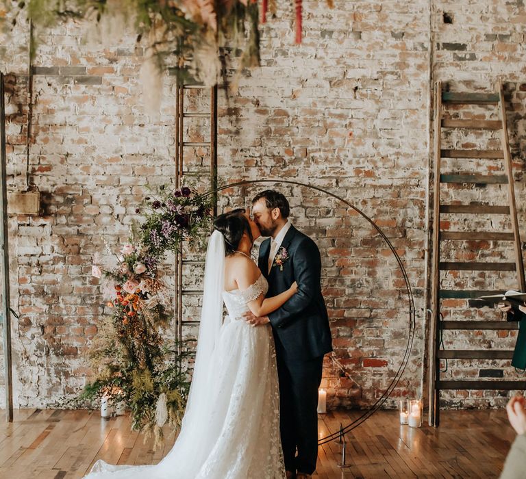 Bride in lace wedding dress and veil kisses groom in blue check suit in front of boho floral moongate with pink, purple and green florals for wedding ceremony in industrial venue