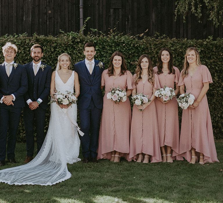 Groomsmen in navy blue three-piece suit and beige tie with bridesmaids in muted pink button up dresses and bride in lace wedding dress