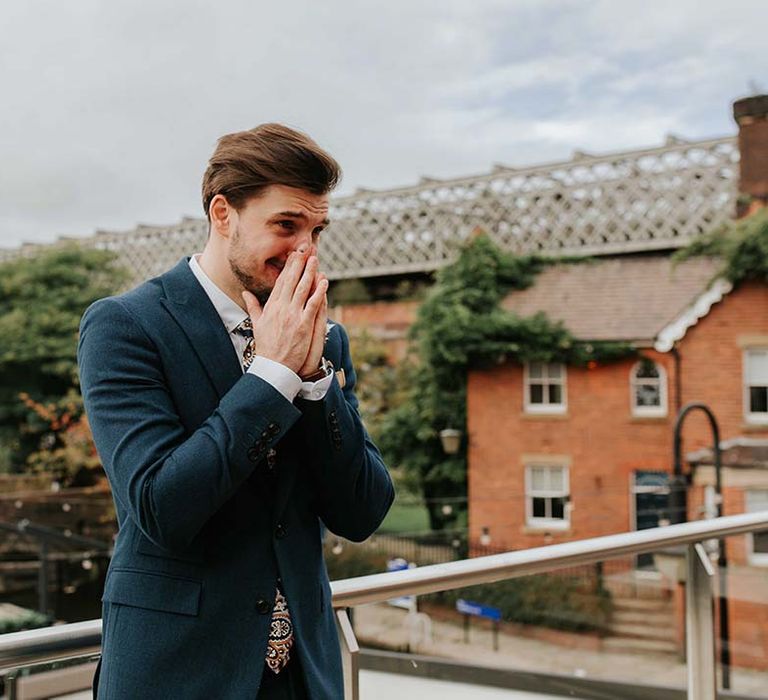 Groom in blue suit and paisley tie puts his hands to his face and tears up as he sees his bride for the very first time