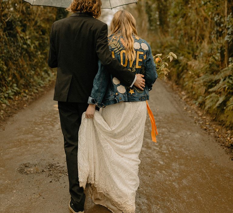 Bride in lace wedding dress and embroidered denim jacket under clear umbrella with groom in black tie