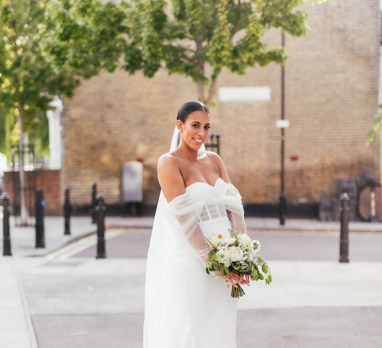 Bride in off the shoulder wedding dress with sheer sleeves and sleek hair with veil holding white flower bouquet 