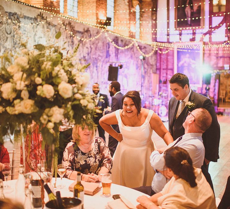 Bride and groom speak to guests at the reception with coloured and fairy lights and tables decorated with white rose centrepieces 