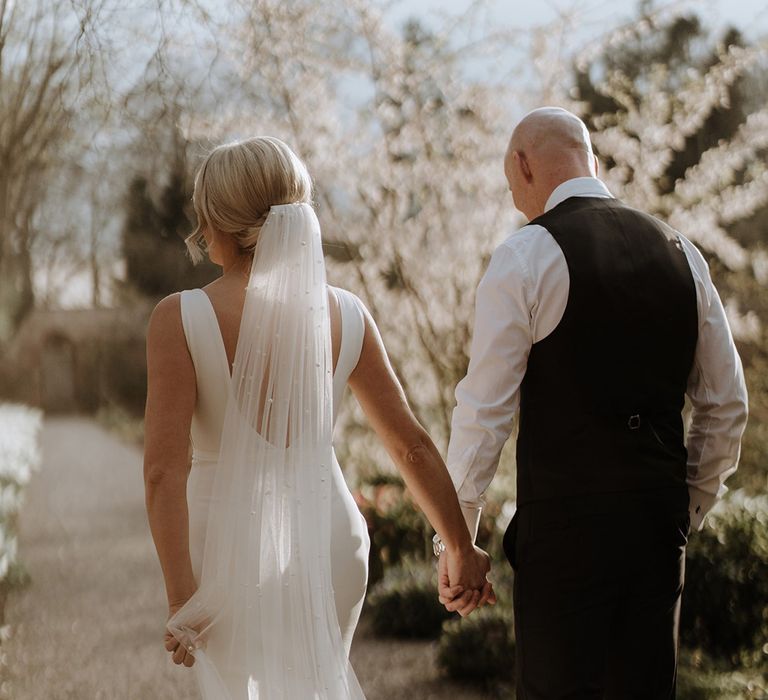 Bride in button back wedding dress and pearl veil holding hands with groom in black trousers and waistcoat 