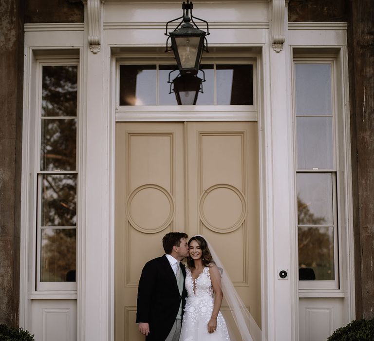 Groom in morning suit kisses bride on the cheek in Mira Zwillinger wedding dress in front of large doorway 