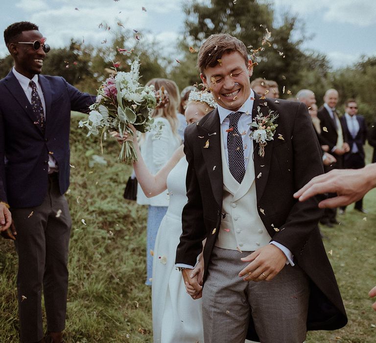 Groom walks through colourful confetti and scrunches his face and holds hands with the bride 