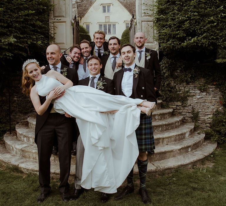 Groom and groomsmen lift the bride as they all pose together in their matching morning suits 