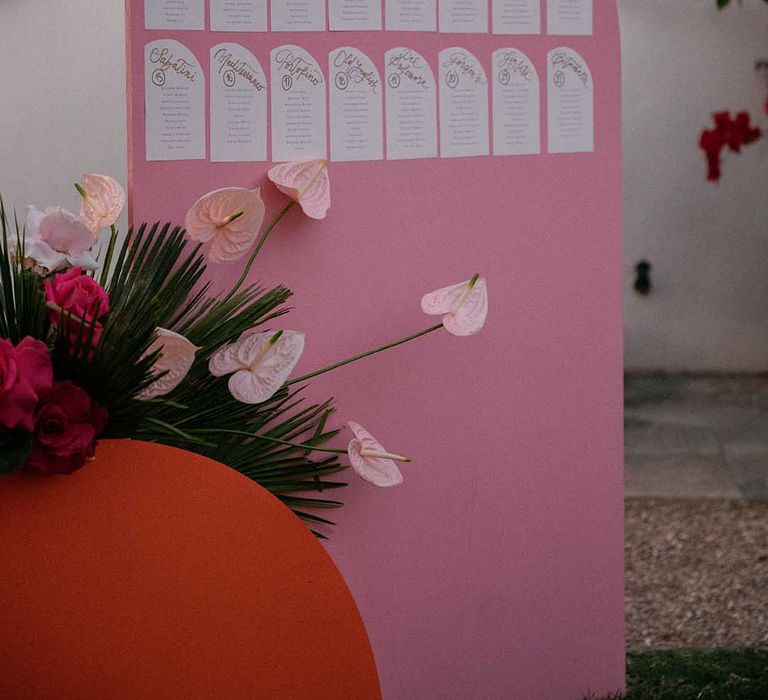 Pink table plan stands behind bright florals and green foliage 