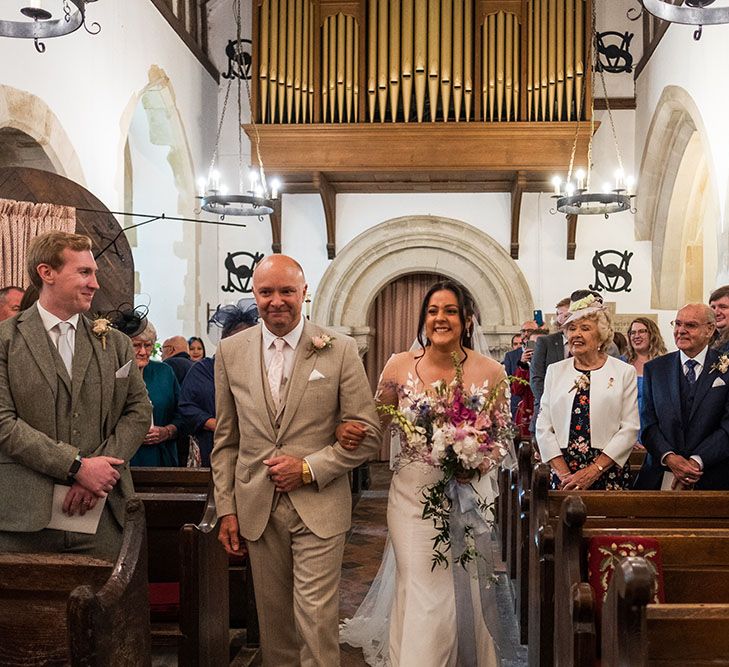 Bride holding colourful tropical bouquet walks down the aisle with her father who wears sand coloured suit 