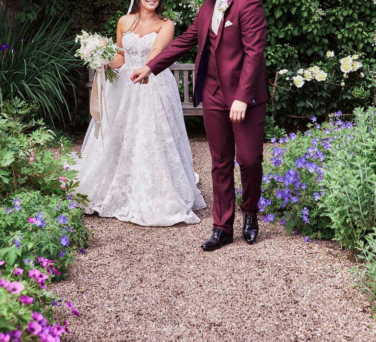 Bride in a strapless lace wedding dress holding hands with there groom in a burgundy coloured wedding suit in the gardens at East Riddlesden Hall