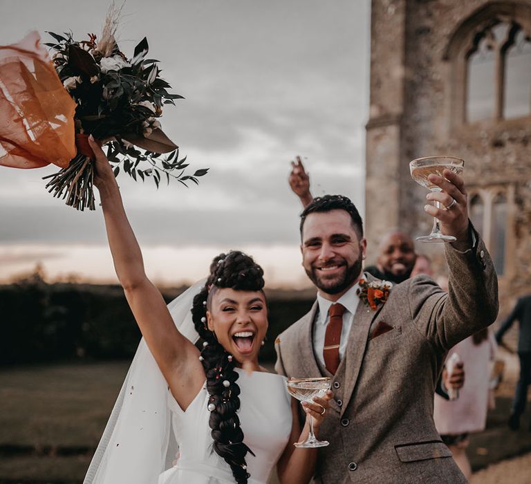 Bride & groom hold champagne glasses as bride lifts her Autumnal bouquet in the air in celebration after Christian wedding ceremony