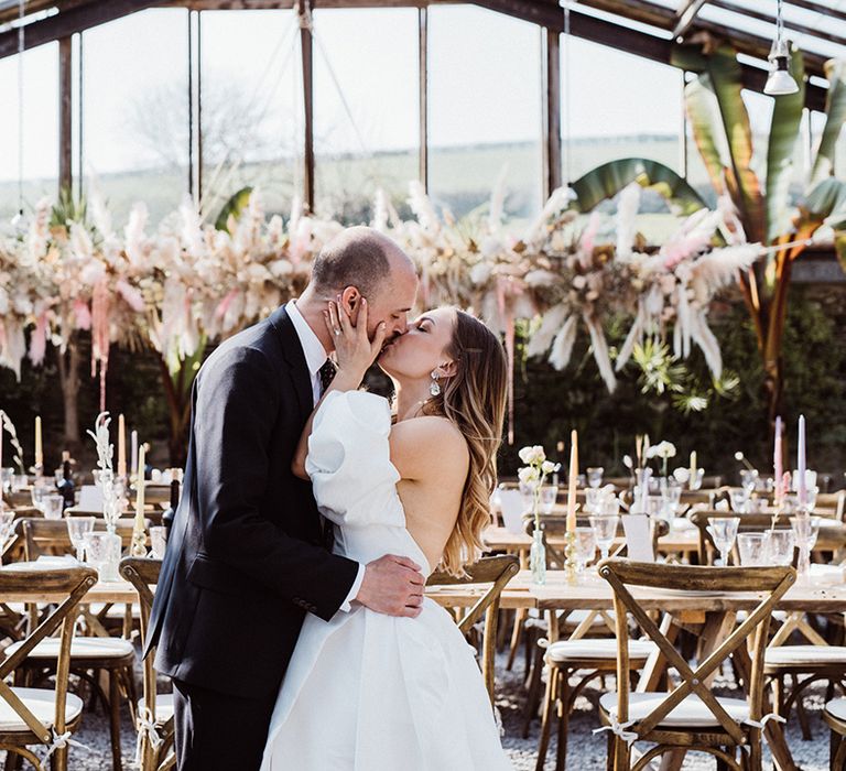 Bride reaches up for the groom and kisses him as they stand in their stunning glasshouse wedding venue 