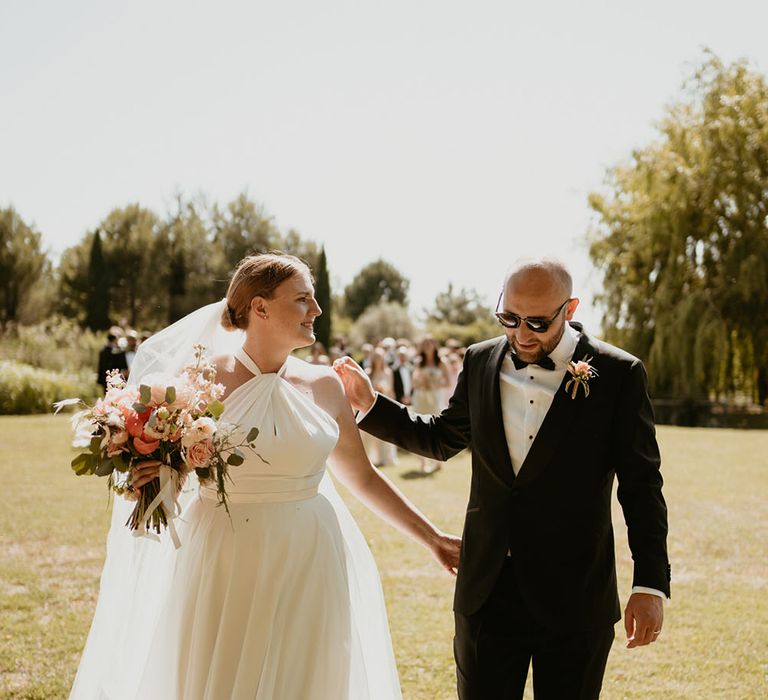 Bride wears her red hair pulled back into low bun whilst carrying peony floral bouquet and walking with her groom in black tie at the Mas Loisonville 