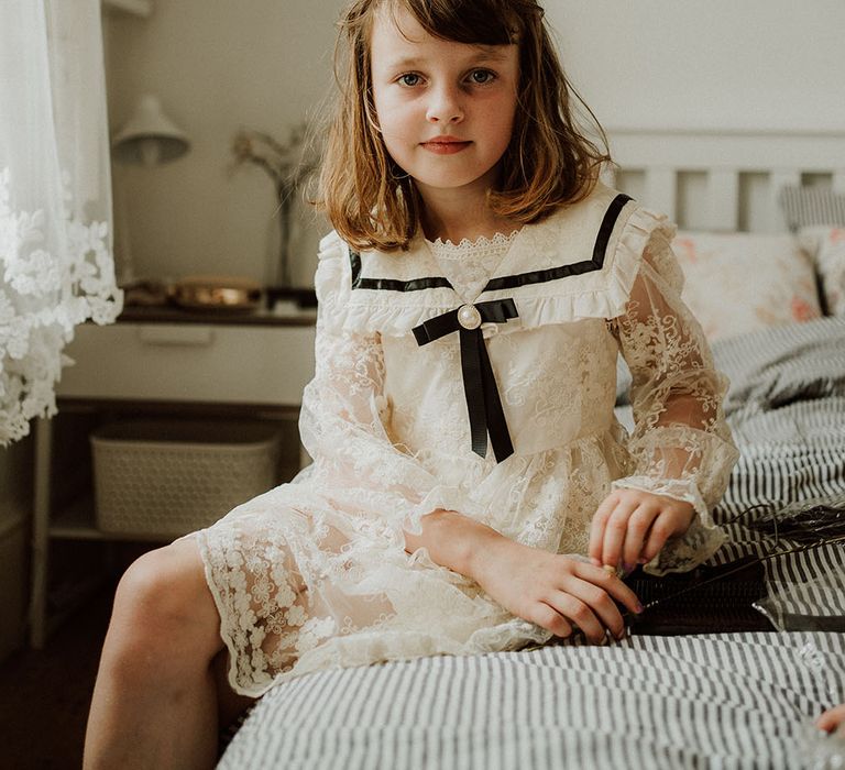 Flower girl wears lace dress complete with black ribbon to the front and sheer sleeves 