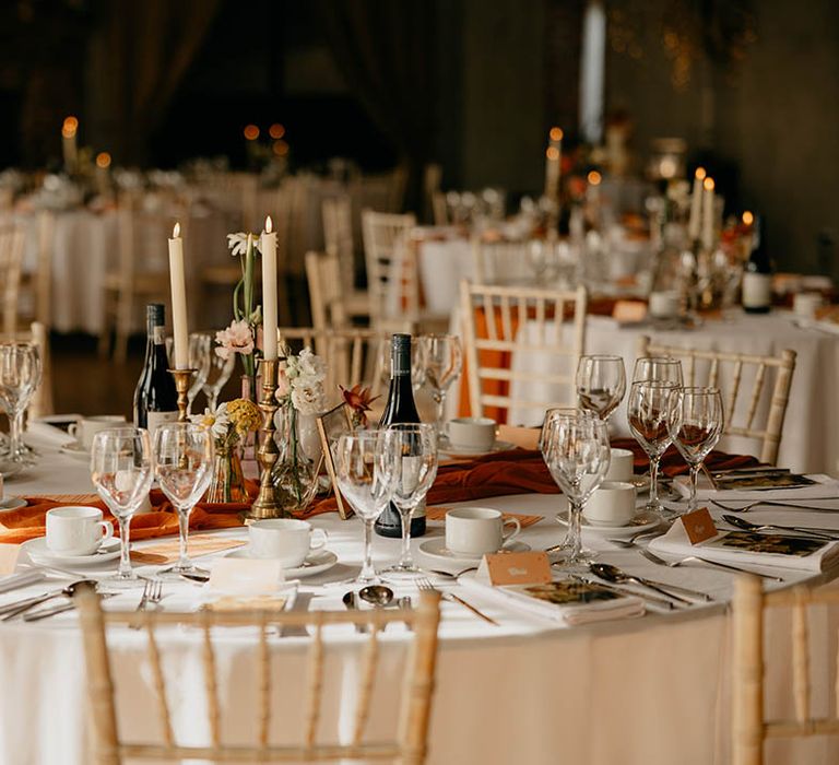 Round white tables with orange runners and white candles in gold candleholders 