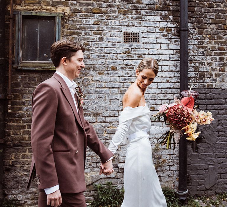 Bride in an off the shoulder wedding dress walking with the groom in a brown suit for their city wedding holding a pink, yellow and brown bouquet 