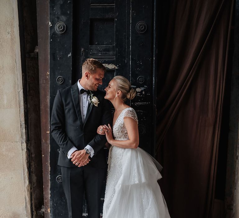 Bride wears capped sleeve embellished wedding dress as stands beside her groom in black tie and floral buttonhole 