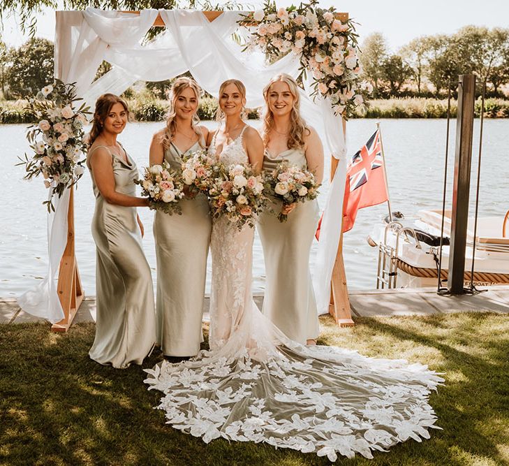 Bride stands with her bridesmaids wearing pastel green satin bridesmaid dresses in front of the Thames for outdoor wedding ceremony