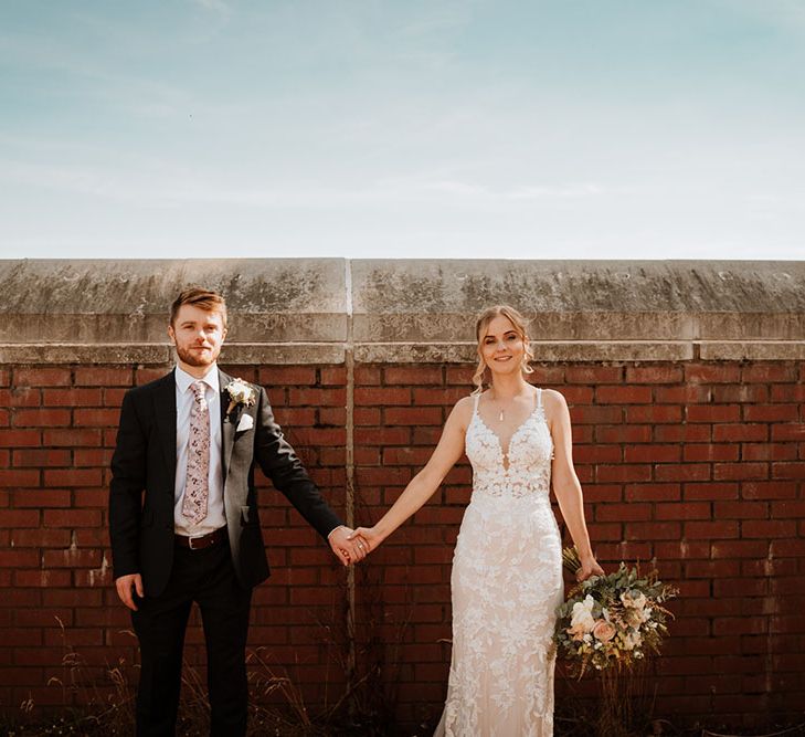 Bride wears lace wedding dress and stands with her groom in front of brick wall for outdoor couples portraits 