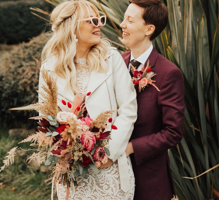 Bride in a lace long sleeve wedding dress with a cream leather jacket and retro cat eye sunglasses with her partner in a burgundy suit 