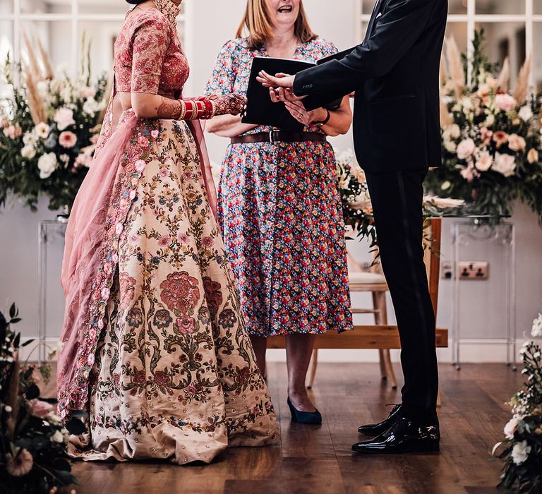 Bride wears beautifully embellished pink lehenga and stands with her groom in black tie during wedding ceremony 