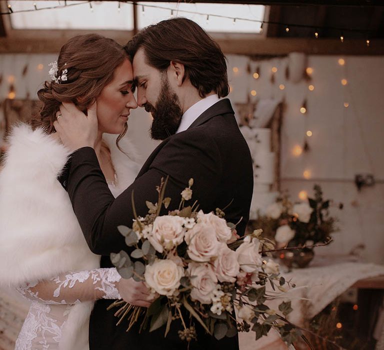 Bride wearing an illusion lace wedding dress and white faux fur shawl rests her forehead against the groom who wears black tie 