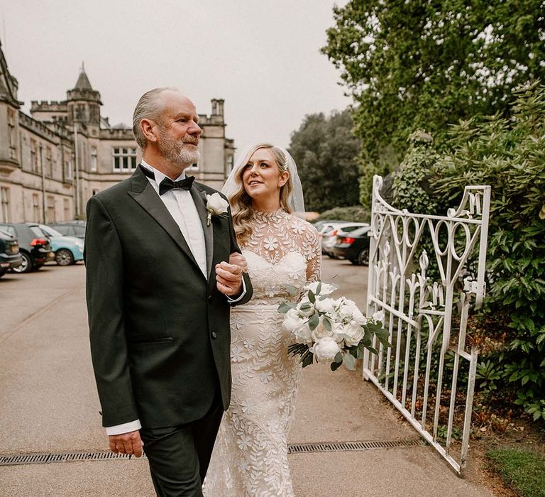 Bride holding white rose bouquet walks with her father wearing black tie 