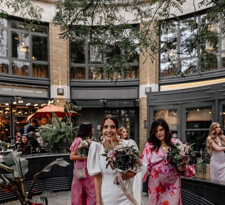 Bride in puff sleeve v neck wedding dress and neutral toned bouquet with her bridal party in mismatched pink bridesmaid dresses