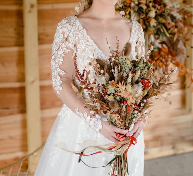 Bride holding dried autumnal wedding bouquet with white dried cotton stems, terra cotta celosia seeds, green foliage wearing long sleeved v neck soft ivory dress with lace floral detailing