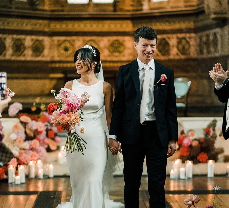 Bride carries bright floral bouquet and walks alongside her groom in black suit and white tie with matching floral buttonhole 