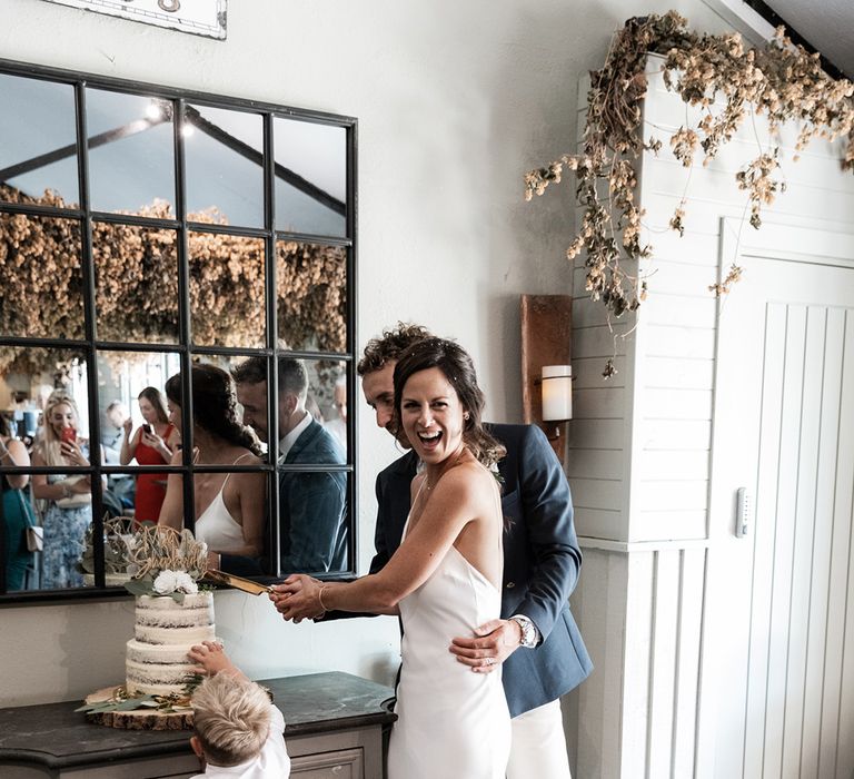 Bride cuts two-tier naked wedding cake with her groom and children at Gate Street Barn