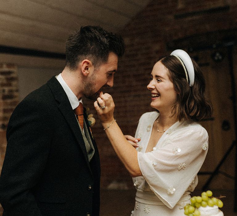 Bride in a pearl flower patterned jumpsuit and headbands feeding the groom some cheese as they cut their wedding cheese tower 