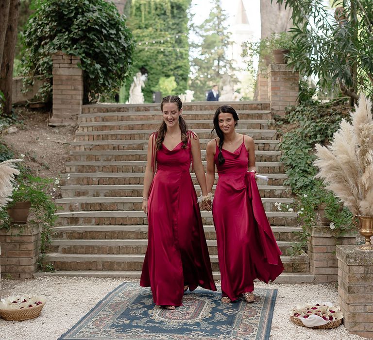 Bridesmaids wear burgundy satin bridesmaid dresses with cowl necks as they walk across Moroccan styled rugs for boho destination wedding