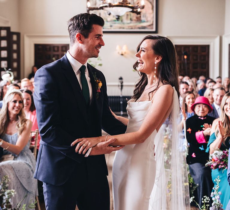 The bride in a sleek cowl neck satin wedding dress with long pearl earrings laughs at the altar with the groom in a navy suit 