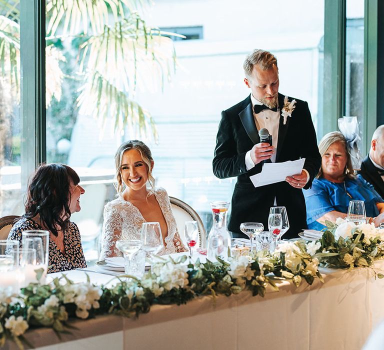 Groom in a black tuxedo standing delivering his wedding speech to the wedding guests and bride 