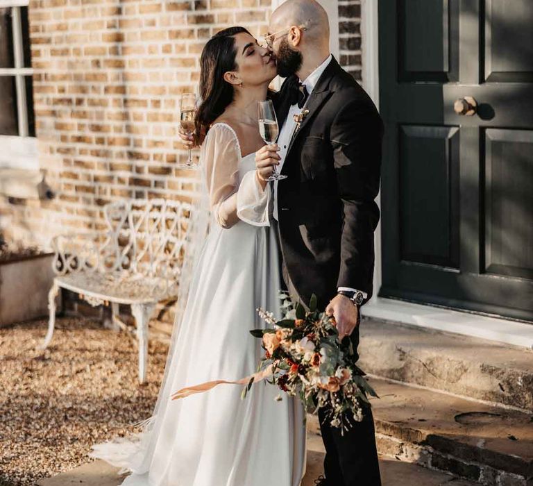 Bride in square neck wedding dress with sheer puff sleeves holding a glass of champagne sharing a kiss with groom in classic black tuxedo with black bowtie 