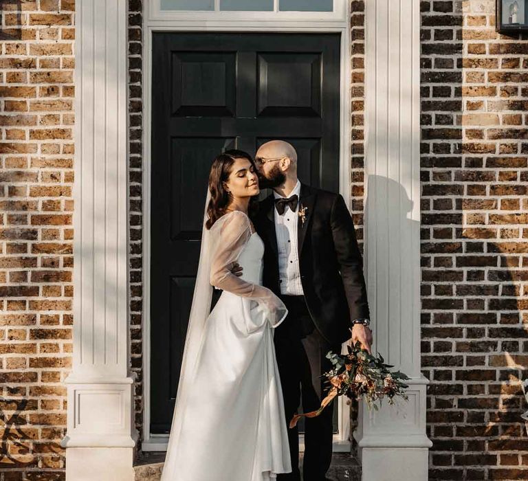 Groom in classic black tuxedo with black bowtie embracing bride in square neck wedding dress with sheer puff sleeves at Elmley Nature Reserve wedding venue 