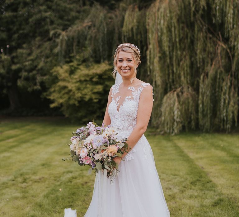 Dando London wedding dress with illusion lace and tulle skirt holding a pastel wedding bouquet with her blonde hair in a braided hairstyle and sparkly headpiece 