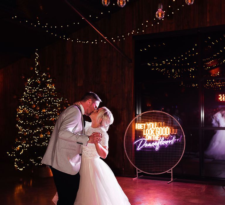 The bride in a tulle bridal skirt dances with the groom on a checkered dance floor with festoon lighting and Christmas tree decoration with neon sign