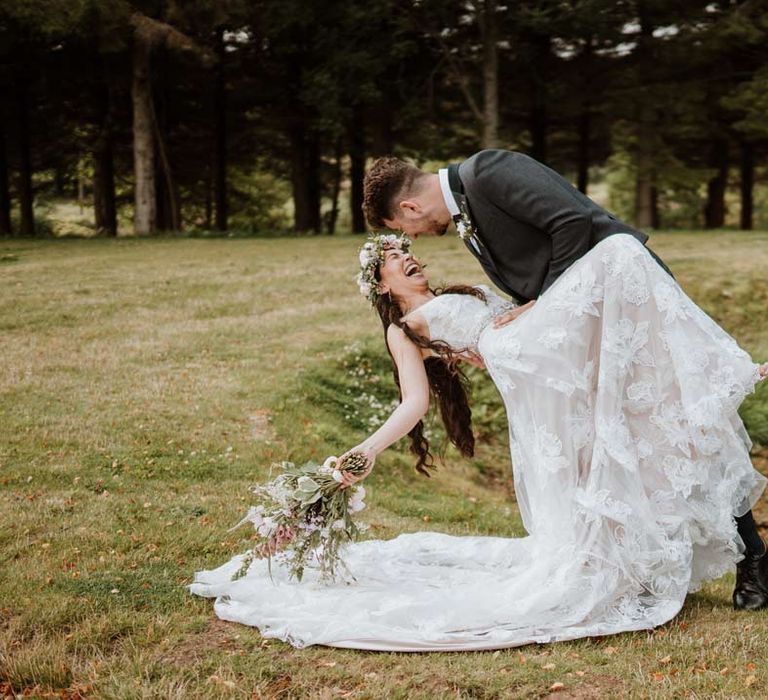 Groom in dark grey blazer dancing and laughing with bride in 3d applique sheer overlay wedding dress with white garden rose flower crown and cathedral-length veil